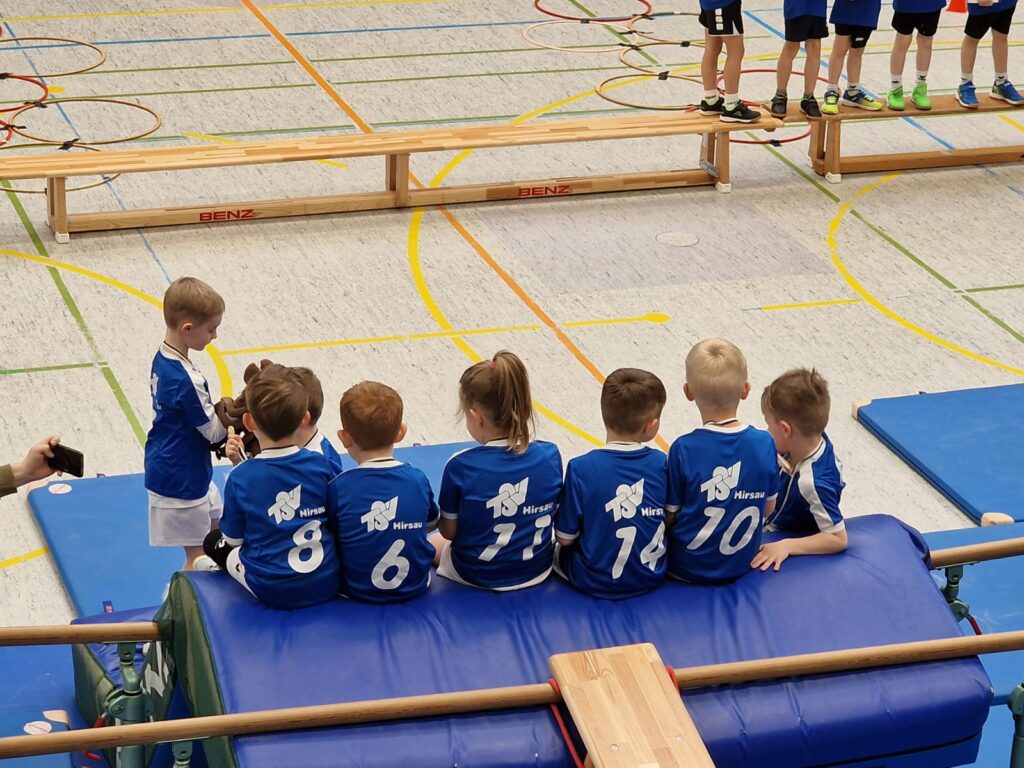 Sechs Kinder des TSV Hirsau sitzen in blauen Trikots bei einem Hallenturnier auf einer Matte, während ein weiteres Kind steht und ein Maskottchen hält.